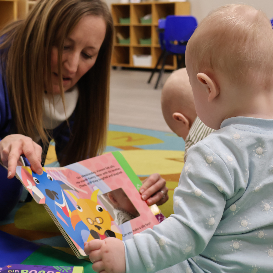Two babies having a board book read to them on the carpet